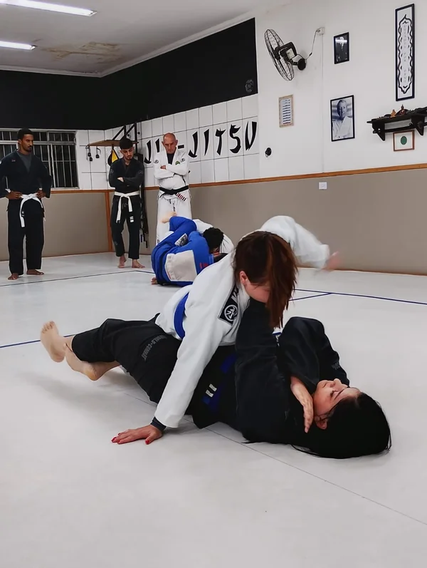 Alunas praticando jiu-jitsu no solo enquanto a turma acompanha